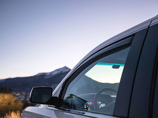 Close-up of a BMW X3 (G01) 2017- with WeatherTech Side Window Deflectors in Dark Tint. Dashboard lights glow softly inside, while mountains and a clear sky are visible through the dusk-lit window and mirror.