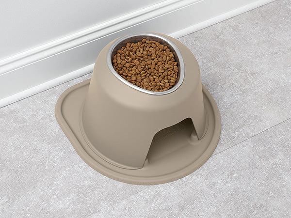 The Weathertech Pair Bowls Stainless Steel pet feeder, in beige, is filled with dry kibble and placed on a tiled floor near a white wall. Its base has a small arched opening and wide tray to catch spilled food.