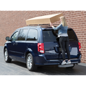 A woman stands on the Weathertech BumpStepXL 24 - AU Version (Standard Plated Hitch Hardware) at the rear of a dark blue minivan, securing a large cardboard box onto the roof rack near a brick wall.