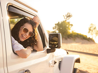 A smiling woman in sunglasses leans out the window of her white BMW X3, enjoying a sunny beach day. WeatherTech Side Window Deflectors Dark Tint help keep the breeze just right as trees and sand create a relaxing backdrop.