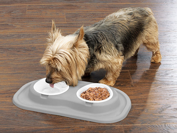 A small dog with long, tan, and gray fur drinks from the WeatherTech Double Compact Pet Feeding System by Weathertech. The dog stands on a wooden floor, and the system’s space-saving bowls hold water and food on a gray mat.