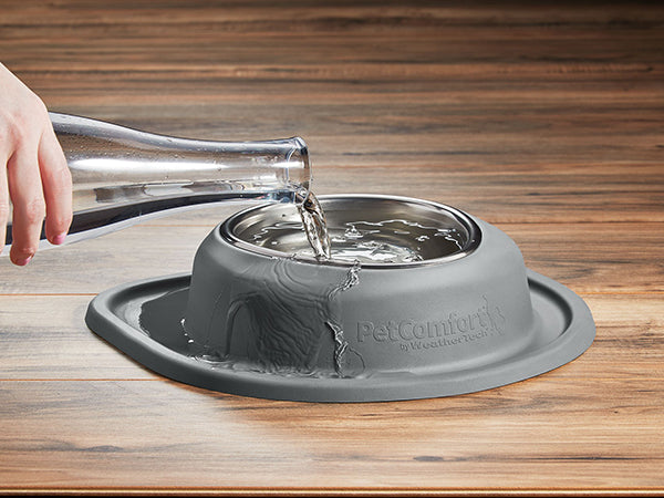 A hand pours water from a glass bottle into a gray WeatherTech Single Compact Pet Feeding System bowl, causing water to overflow onto the surrounding mat on a wooden surface.