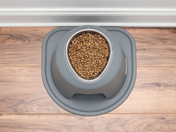 The WeatherTech Single Elevated Pet Feeding System by Weathertech, in gray, holds dry kibble and rests on a matching silicone mat on a wooden floor near a white baseboard.