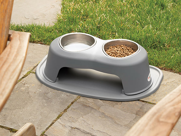 The WeatherTech Double Elevated Pet Feeding System by Weathertech, featuring two stainless steel bowls in a gray holder, sits on a stone patio—one bowl holds dry kibble while the other is empty. Grass is visible in the background.