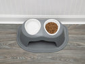 The WeatherTech Single Pet Feeding Bowl Plastic by Weathertech sits on a wood-patterned floor near a white paneled wall, with one of its non-toxic bowls filled with brown kibble and the other left empty.