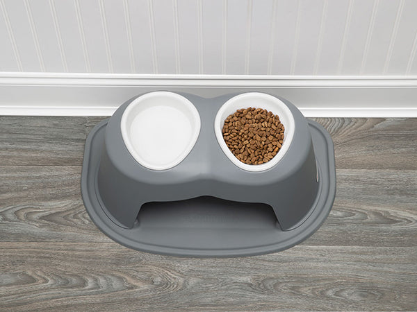 The WeatherTech Single Pet Feeding Bowl Plastic by Weathertech sits on a wood-patterned floor near a white paneled wall, with one of its non-toxic bowls filled with brown kibble and the other left empty.