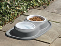 The WeatherTech Double Compact Pet Feeding System by Weathertech is an ergonomic pet bowl with separate sides for water and kibble, shown on a stone patio surrounded by lush green ivy.