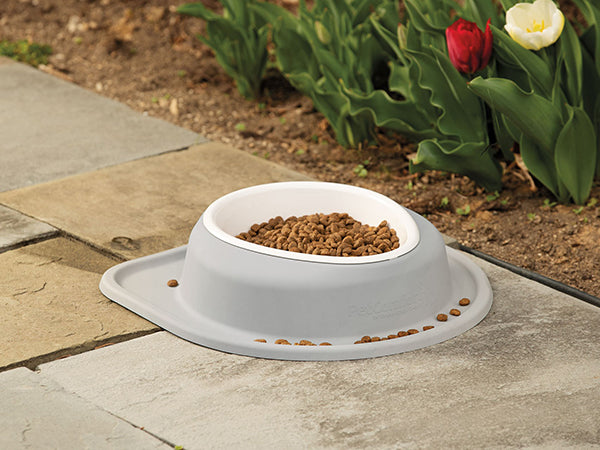 The WeatherTech Single Compact Pet Feeding System by WeatherTech is a white ergonomic dog bowl filled with brown kibble, placed on a stone patio near blooming red and white tulips, with some kibble scattered around.