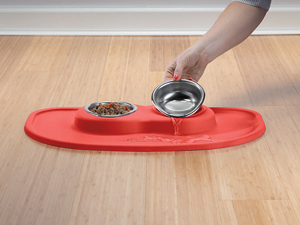A hand pours water into a metal bowl in the WeatherTech Double Compact Pet Feeding System, featuring a red silicone mat on a wooden floor. An ergonomic kibble bowl sits beside it—this space-saving feeder by WeatherTech fits any home.