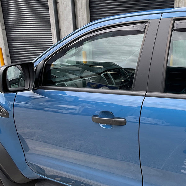 A blue car with WeatherTech Side Window Deflectors Dark Tint (for Ford Ranger PX2/PX3 and Mazda BT-50 2012-2020) is parked by a building with black roller doors, showing the drivers side and its reflection in the window channel.