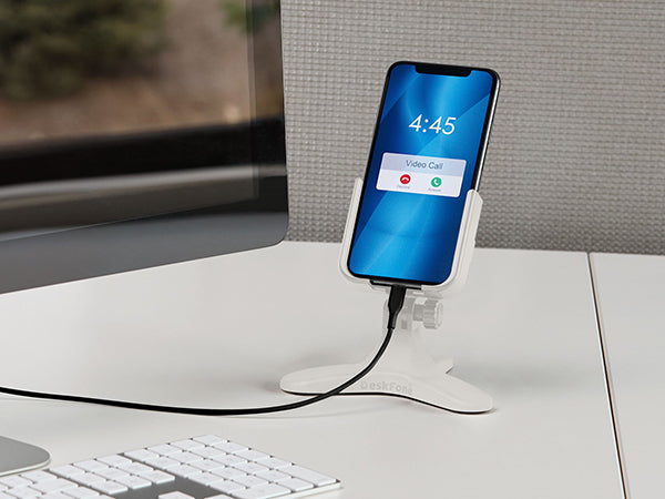 A smartphone rests on a WeatherTech DeskFone White adjustable phone stand, plugged in and displaying an incoming video call. It’s positioned on a desk next to a keyboard and monitor.