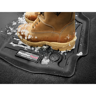 A pair of tan boots with snow on them stands on a black WeatherTech FloorLiner HP for Jeep Wrangler (2018-) 1st Row, providing superior protection as snow and ice cover its surface.