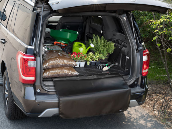 The open trunk of a MINI Countryman displays gardening supplies, including soil, potted flowers, and herbs, all neatly arranged on a WeatherTech CargoLiner - MINI Countryman (U25) - Black, with trees and grass visible beside the vehicle.
