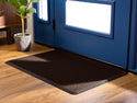 A large, dark brown WeatherTech Carpet Mat by Weathertech rests on wood flooring in front of a blue door beside a potted plant, as sunlight streams through the door’s lower window panels and brightens the entryway.