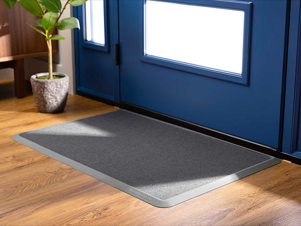 The WeatherTech Carpet Mat by Weathertech, in gray with a light border, rests on a wooden floor before blue double doors with glass panels. A potted plant is nearby as sunlight streams through the doors.