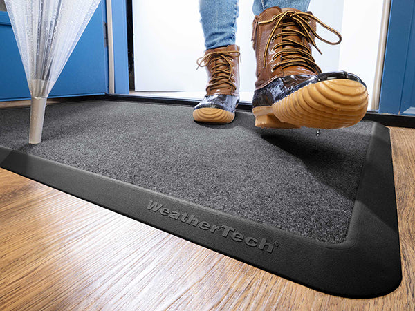 A person in wet boots steps onto a Weathertech Carpet Mat inside a doorway, while an umbrella rests nearby on the wooden floor.