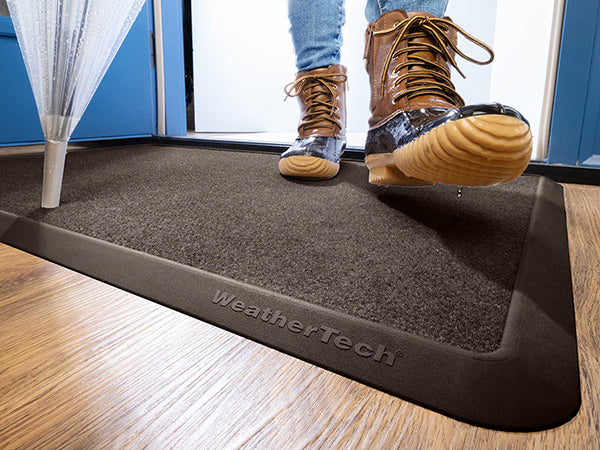 A person in brown boots steps onto a dark WeatherTech Carpet Mat by Weathertech at an entryway, with an umbrella nearby and water dripping from their boot.