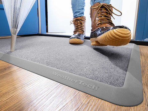 A person wearing brown duck boots steps onto a WeatherTech Carpet Mat by WeatherTech inside a doorway; water drips from their boots and umbrella, keeping the floor dry.