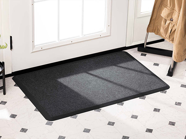 A WeatherTech Carpet Mat in dark gray from Weathertech is laid inside a bright entryway with white walls and tile flooring. Sunlight pours in through glass panels, and a tan coat hangs on a black rack by the door.