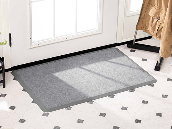 A WeatherTech Carpet Mat by WeatherTech in light gray sits on a white tiled floor with black diamond accents near a glass door; a coat hangs on a nearby rack.