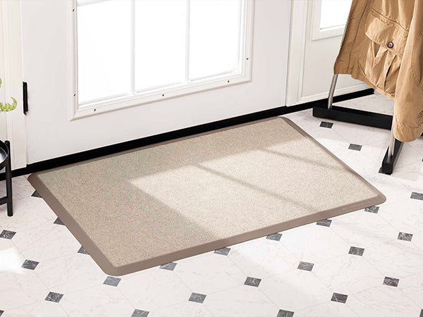 The WeatherTech Carpet Mat by WeatherTech rests on a white tile floor with black diamond accents near a white door with glass panes, while sunlight filters in and a tan coat hangs on a nearby rack.