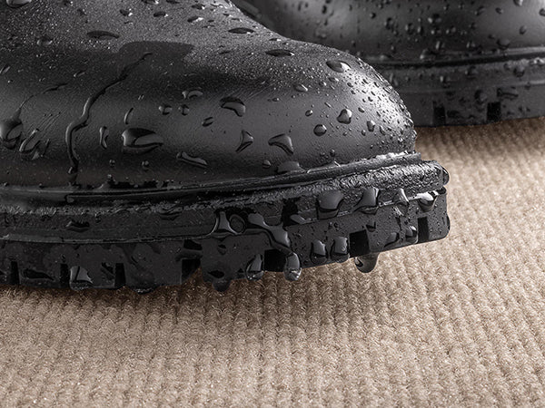 A black boot covered in water droplets rests on a WeatherTech Carpet Mat by Weathertech, with water dripping from the sole onto the mat’s light brown, commercial-grade textured surface.