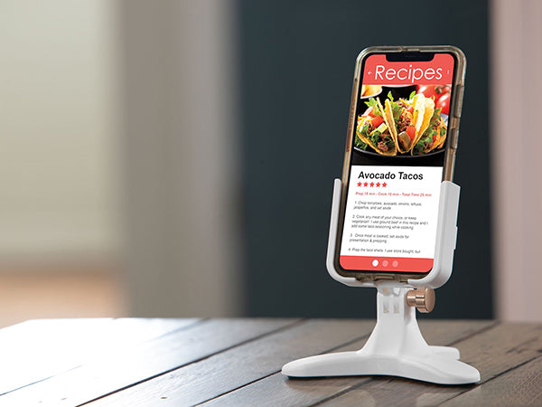 A Weathertech DeskFone White holds a smartphone on a wooden table indoors, displaying an avocado taco recipe with photos and ingredients—making for a convenient desktop phone holder setup.