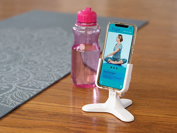 A WeatherTech DeskFone White holds a smartphone showing a yoga app with a woman seated, beside a pink water bottle and a gray yoga mat on the wooden floor.