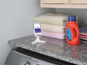 A Weathertech DeskFone White holds a smartphone displaying a timer on a granite countertop beside folded towels and liquid laundry detergent, positioned above a washing machine.