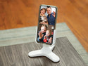 A Weathertech DeskFone White holds a smartphone displaying a video call with two adults, a child, and a dog smiling and waving, set on a wooden table with a blurred wood floor background.
