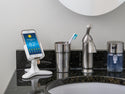 A smartphone showing Chicagos weather rests on a Weathertech DeskFone White adjustable stand on a bathroom counter beside a toothbrush in a cup, soap dispenser, and sink with a mirror above.