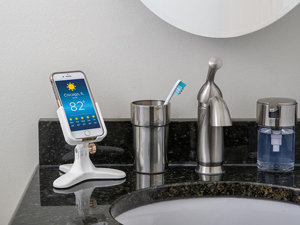 A smartphone showing Chicagos weather rests on a Weathertech DeskFone White adjustable stand on a bathroom counter beside a toothbrush in a cup, soap dispenser, and sink with a mirror above.